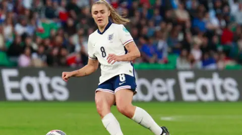 PA Media Georgia Stanway playing football in the blue and white England kit. She has long, light hair tied back in a ponytail and is looking down at the ball in front of her.