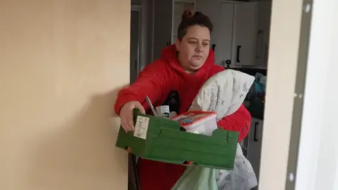 Qays Najm/BBC A woman in a red fleece carries a green cardboard banana box containing small items as she moves through a doorway, with a pillow under her left arm.