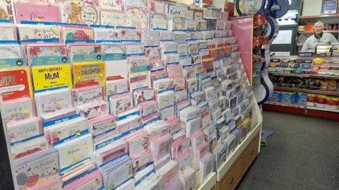 Gordon Bird A huge wrack of birthday cards in a newsagents where an elderly man in a grey cardigan stands behind a confectionary counter