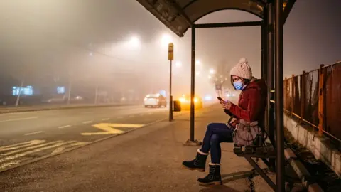 Getty Images woman wearing mask waits at a bus stop on a foggy night