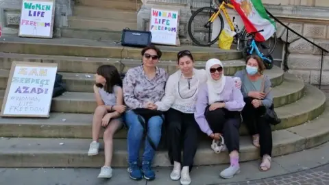 Cad Taylor/BBC Four women and a girl sit on the steps of Ipswich Town Hall during a vigil.