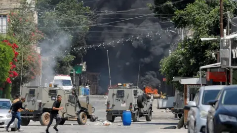 Reuters Palestinians run for cover as Israeli military vehicles move through Jenin, in the occupied West Bank (3 July 2023)