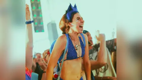 Alastair Brookes A festivalgoer wears a blue bikini top with matching blue headband with cat ears. They have stars stuck to their face, with their mouth open as if in song. People are partying around them and all seemingly facing towards a stage that is not in shot. 