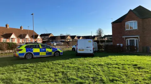 BBC A police car and two white vans parked on grass next to a house. A row of houses can be seen in the background. The sky is blue.