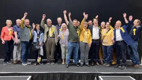 The Liberal Democrat group lines up after the votes are counted in a celebratory photograph. Many have their arms in the air showing their happiness at the outcome and they are wearing yellow rosettes.