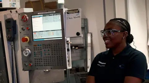 In-Comm Training A woman wearing protective glasses standings next to an electronic machine with a screen and dozens of buttons. It appears to be inside a factory or warehouse.
