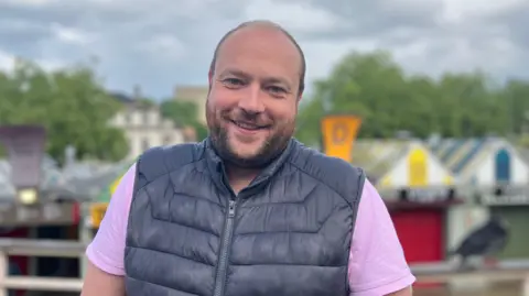 Owen Sennitt/BBC A man wearing a pink shirt under a navy blue gilet smiles towards the camera, with colourful market stalls out of focus behind him