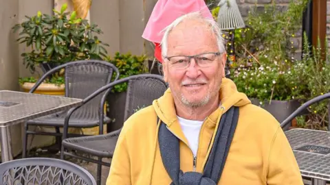 Larry Gibbons smiles at the camera as he sits in the seating area outside his Largigi cafe. He is wearing a yellow hoody and glasses.