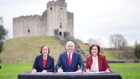 PA Media Secretary of State for Wales Jo Stevens, Defence Secretary John Healey and First Minister of Wales Eluned Morgan during a visit to Cardiff Castle which is in the background. They sit at a table with documents in front of them as they hold pens preparing to sign a defence deal.