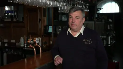 Matthew Russell stands at the bar of the Pumproom in Chatham. He wears a black logoed sweater and a white shirt.