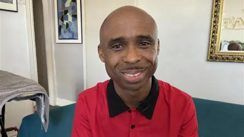Ikenna Ajiero wearing a red shirt and sat on a sofa, smiling.