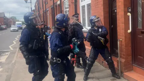 BBC Photograph of police on a drugs raid in Wigan. The image shows police officers in riot gear smashing down the front door of a house in the Scholes area of the town.