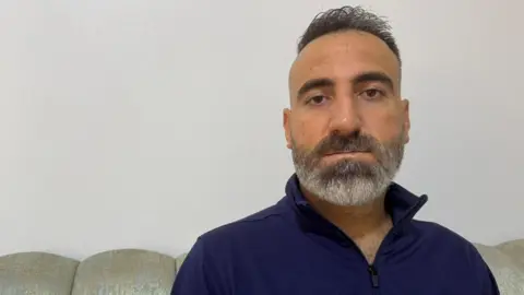 A man in his 40s with a greying beard and short dark hair looks directly at the camera 