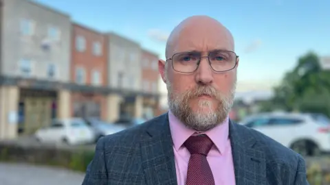 Troy Smith looking at the camera. He has a beard and is wearing a dark grey suit jacket, pink shirt, maroon tie and thin-framed glasses