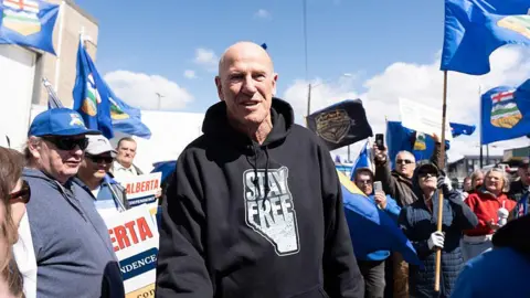 AFP via Getty Images Separatist leader Mitch Sylvestre is seen during a rally in front of the Elections Alberta headquarters in Edmonton. He is wearing a black hoodie with an Alberta map logo that says STAY FREE. He is an older, bald gentleman. Behind him are people waving Alberta flags and holding signs that say ALBERTA INDEPENDENCE.