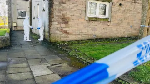 BBC Two people in white forensic suits and blue gloves, outside a building, with blue and white police tape in the foreground.