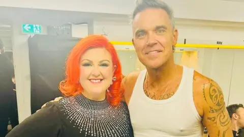 Michelle McManus with vibrant red hair and a black dress with diamantes standing next to Robbie Williams, who has grey hair spiked up and wearing a white vest. He has lots of tattoos. They are pictured ahead of their performance at Murrayfield stadium.