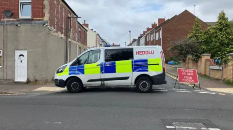 A police van on a road. There is a sign in red that reads road closed, in white font. Which is translated in Welsh. There is a house to the left of the image with a white door.
