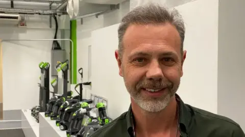 A man with grey hair and a beard, wearing a dark green shirt is on the right of the photo smiling at the camera. A number of vacuum cleaners are on the left in the background.