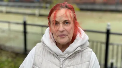 BBC A woman with red hair, wearing a white padded coat, is standing in front of black railings that run alongside a river bank.