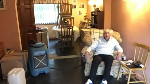 BBC A man sits in a chair in a room with a large fan and chairs on tables