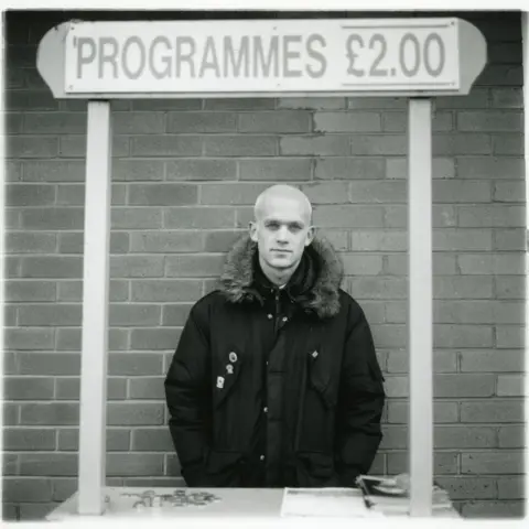 Jason Scott Tilley/Lower Block A short-haired blonde programme seller stands below a sign stating programmes were being sold for £2. He is wearing a dark snorkel parka with some badges.