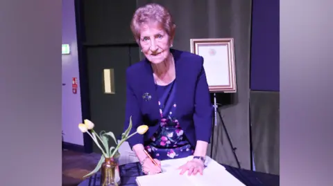 Dame Norma Redfearn, with short brown/grey hair, wears a navy dress suit. The dress is also navy with pink patterned flowers on it. She is signing a book which is on a table with a tartan tablecloth yellow tulips.