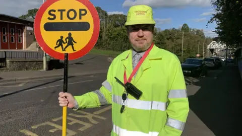 Tony Young outside Crumlin Integrated Primary School wearing shirt and tie and high vis jacket, with high vis hat that reads "crossing patrol" and lollipop stick sign with the word STOP on it