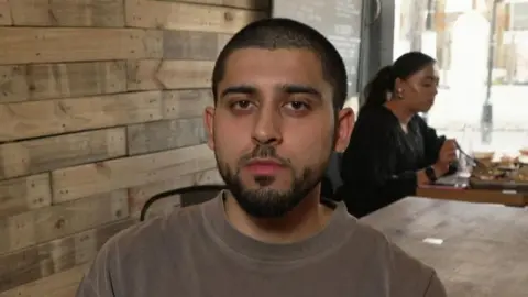 A man sits at a wooden table inside a café or restaurant. Other customers are seated at a table by a window in the background.