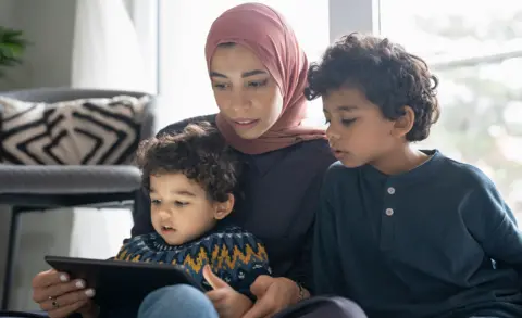 Getty Images Stock image shows a mother holding a tablet while watching something on the screen with two young children at home in a living room.