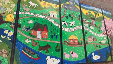 A large colourful mural depicting farmyard animals, the school, a river, swans, and a church. There is a scrawling ribbon across the whole thing on which reads "over all these things put on love, which binds everything together in perfect harmony." 