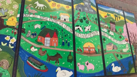 A large colourful mural depicting farmyard animals, the school, a river, swans, and a church. There is a scrawling ribbon across the whole thing on which reads "over all these things put on love, which binds everything together in perfect harmony." 