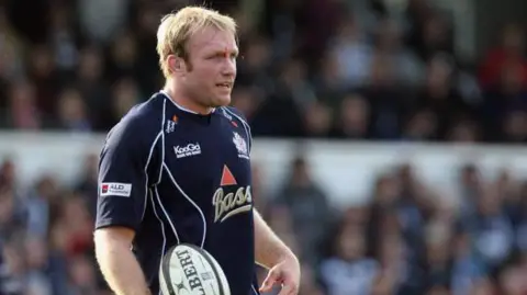 Matt Cardy Matt Salter during Guiness Premiership Match. He's wearing dark blue kit with gold writing and red and white logos. He has blonde hair and a cut next to his right eyebrow. He's holding a black and white Gilbert rugby ball and looking to the distance.