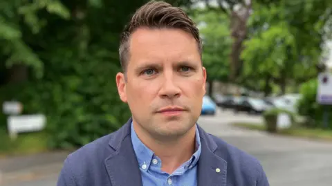 Olivia Richwald/BBC A man with short brown hair wearing a blue shirt and navy jacket stands in a carpark