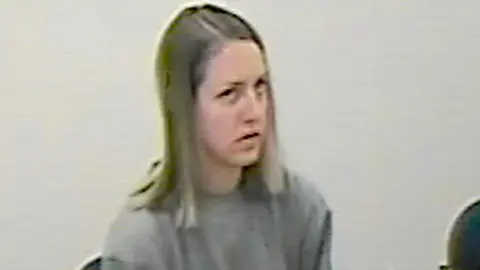 CCTV footage of Lucy Letby, who has shoulder length blonde hair and wears a plain grey jumper, sitting in a police interview room.