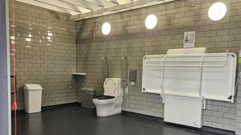 Liz Owen Inside the new changing space, which is a large room with brick shaped sage coloured tiles and a dark grey laminate flooring. There are three circle shaped lights on the right wall of the rectangle shaped room, just above a large pull down table. There is a toilet, bin and red emergency chord against the wall. 