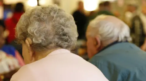 A stock image of two older people, sat with their backs to the camera. An elderly woman is in the foreground of the image. She has short grey curly hair and wears a pale pink jumper and hearing aid. An older man sits in the background. He has grey hair and wears a green jacket, as well as a hearing aid. 