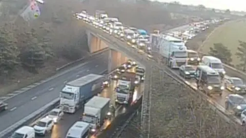 National Highways A traffic camera shows the A14 and a bridge carrying the A11 over the top of it. There are queues of vehicles on both roads. Most vehicles have their headlights on.
