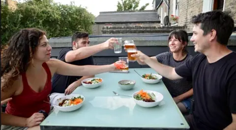 Two couples clink glasses and sit at a table eating food in the garden. The two men wear dark t-shirts. One woman wears a maroon top and the other wears a stripy T-shirt.