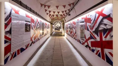 BBC An exhibit inside Jersey War Tunnels. The displays have Union and Jersey flags dotted along them along with information boards. Union flag bunting is hanging from the tunnel's ceiling.