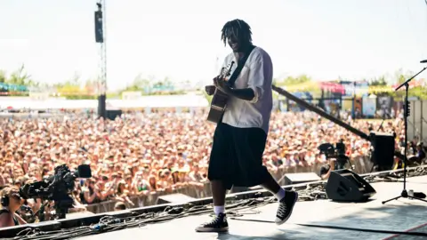Myles Smith wearing a white top with rolled-up sleeves and oversized black shorts smiles while he plays guitar on stage in front of a huge crowd at Radio 1's Big Weekend in Sunderland.