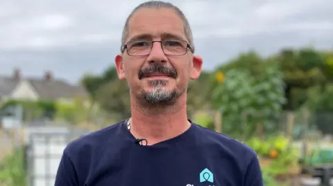 Ricky smiling at the camera. He has shaved grey hair, a moustache and goatee beard. He wears glasses and a navy blue t shirt. He is standing in an allotment with trees in the distance.