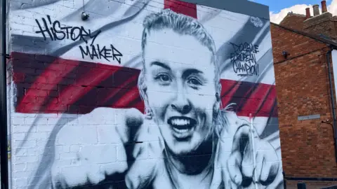 Justin Dealey/BBC A large painting of Leah Williamson pointing with both hands is on the side of a brick wall. She is backed by the white and red England flat.
