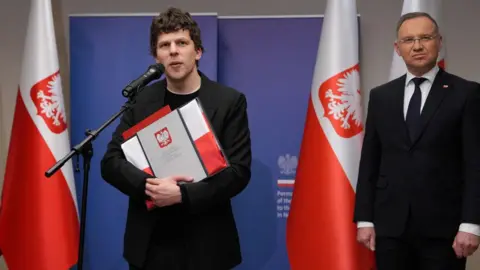 Reuters Jesse Eisenberg standing holding a folder with Polish colours and emblem, speaking into a microphone flanked by two large Polish flags and President Andrzej Duda