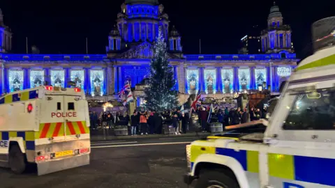 A blue lit Belfast City Hall. There is a Christmas tree in front and people carrying Union Jack flags have gathered at the gates. Two police land rovers are on the road. 