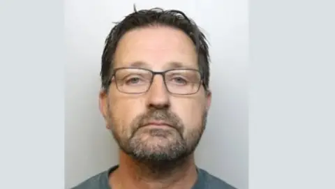 A police custody mugshot of Kevin Thompson, who has a beard and short hair and is wearing glasses