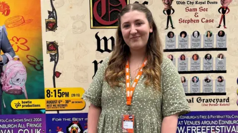 A woman in a long green dress wearing an orange lanyward standings in front of a wall of posters for Edinburgh festival shows
