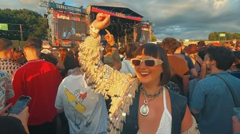 Lucy Smith-Butler Lucy stands in a crowd in front of a stage which says Truck Festival on it, surrounded by people looking away from camera at the stage as she looks towards it, smiling with her arm in the air. She has short brown hair with a fringe over her forehead, and is wearing a sparkly tasseled jacket.