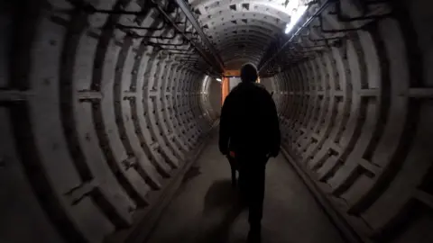 A man pushes a wheelbarrow down the cylindrical entrance to a former nuclear bunker in Edinburgh. You can see his silhouette as he walks down the corridor towards a doorway in the distance.