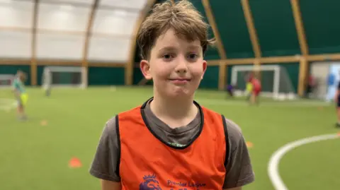 Noah (11) dengan rambut coklat pendek berdiri di tepi lapangan sepak bola dalam ruangan mengenakan bib Premier League Kicks berwarna oranye terang.